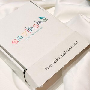 Puede incluir: Una caja de env&iacute;o de cart&oacute;n blanco con el logotipo colorido "envikshe Handcrafted Accessories" y un peque&ntilde;o icono de p&aacute;jaro. La solapa de la caja tiene impreso el texto negro "Your order made our day!" en una fuente sans-serif clara.