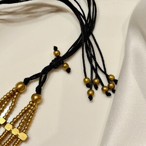 Puede incluir: Un collar que presenta m&uacute;ltiples hileras de peque&ntilde;as cuentas redondas de color dorado met&aacute;lico y espaciadores rectangulares dorados. Las hileras est&aacute;n unidas por un cord&oacute;n negro con un nudo corredizo ajustable, terminado con peque&ntilde;as cuentas doradas en los extremos.