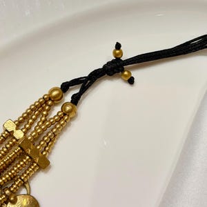 Puede incluir: Un collar de m&uacute;ltiples hilos con filas de peque&ntilde;as cuentas doradas mate y espaciadores rectangulares dorados. Los hilos se unen mediante un cord&oacute;n negro con un nudo corredizo, decorado con dos peque&ntilde;as cuentas doradas en el cierre.