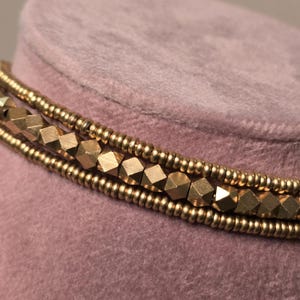 May include: Close-up of a gold-colored beaded necklace with multiple strands. The necklace features small, round beads and larger, geometric-shaped beads. The background is a soft, muted pink.
