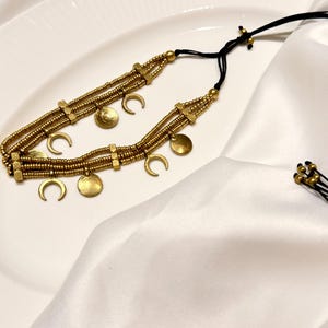 Puede incluir: Un collar de varias hileras con cuentas de tono dorado y dijes met&aacute;licos en forma de lunas crecientes y peque&ntilde;os discos circulares. El collar est&aacute; ensartado en un cord&oacute;n negro fino con nudos deslizantes ajustables y cuentas doradas.