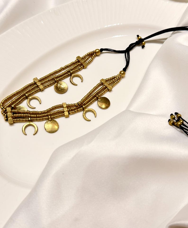 Puede incluir: Un collar de varias hileras con cuentas de tono dorado y dijes met&aacute;licos en forma de lunas crecientes y peque&ntilde;os discos circulares. El collar est&aacute; ensartado en un cord&oacute;n negro fino con nudos deslizantes ajustables y cuentas doradas.