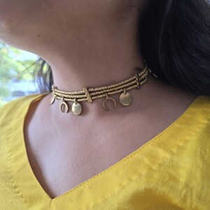 Puede incluir: Un collar tipo gargantilla de tono dorado con tres filas horizontales de peque&ntilde;as cuentas cil&iacute;ndricas texturizadas. El collar est&aacute; adornado con dijes colgantes alternos en forma de lunas crecientes y peque&ntilde;os discos circulares planos.