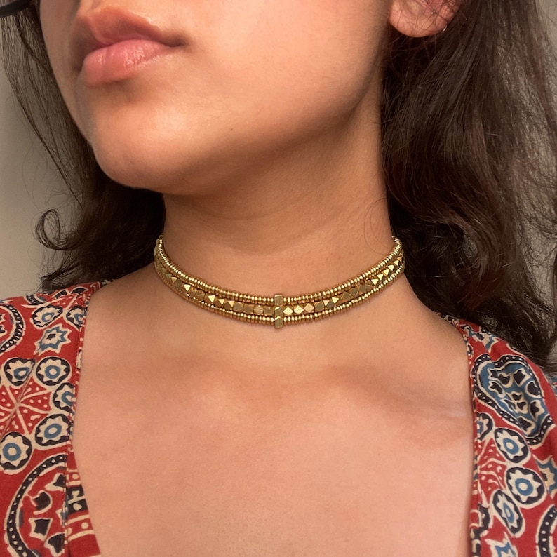 May include: A gold-colored choker necklace with a geometric design. The necklace features small, round beads and square and rectangular accents. The choker is worn around the neck, and the person is wearing a red patterned top.