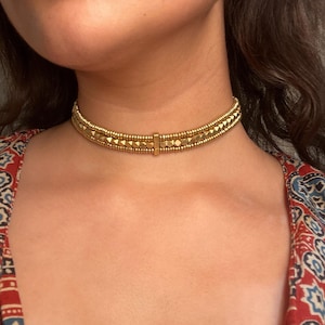 May include: A gold-colored choker necklace with a geometric design. The necklace features small, square-shaped beads and a central rectangular clasp. The choker is worn around the neck, and the person is wearing a patterned top.