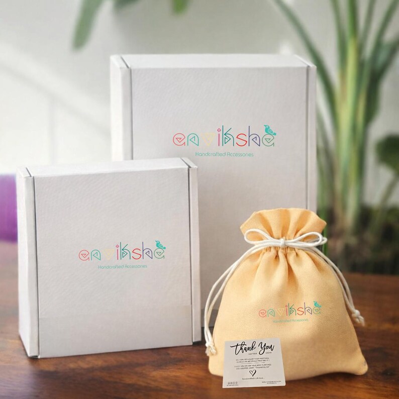 Puede incluir: Dos cajas de cart&oacute;n blanco y una bolsa de tela de color amarillo claro con cord&oacute;n, cada una con el logotipo colorido y el texto "enviksha Handcrafted Accessories". Una peque&ntilde;a tarjeta cuadrada blanca con el texto "Thank You" est&aacute; unida a la bolsa.