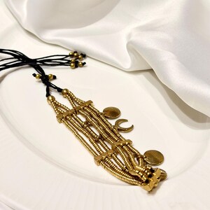 Puede incluir: Una pulsera de varias hileras con cuentas cil&iacute;ndricas de tono dorado met&aacute;lico dispuestas en cuatro filas paralelas, unidas por espaciadores cuadrados. La pieza incluye dijes colgantes circulares y en forma de media luna con cordones negros.