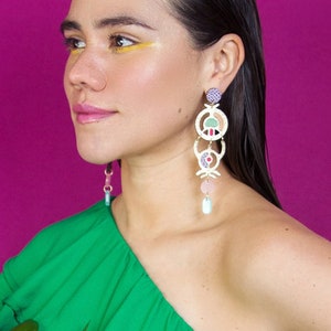Peut inclure: Une femme portant un haut vert à une épaule avec de grandes boucles d'oreilles géométriques colorées. Les boucles d'oreilles sont en métal blanc et ont des accents roses, verts et violets.