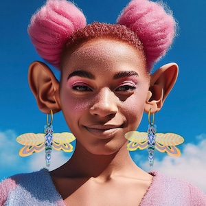 Può includere: Una persona con i capelli rosa in due chignon indossa grandi orecchini a cerchio con ciondoli a forma di libellula. Gli orecchini sono gialli, verdi e blu.