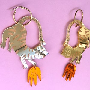Puede incluir: Pendientes de tono dorado con diseño de tigre y una llama naranja colgante. El tigre está representado en una pose juguetona, con la cola enroscada alrededor de su cuerpo.