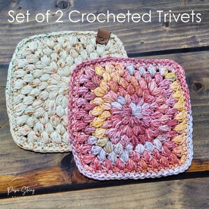 May include: Two crocheted trivets. One is cream-colored with a brown leather loop, the other is a square with a colorful pattern of pink, yellow, and purple. The text "Set of 2 Crocheted Trivets" is at the top.