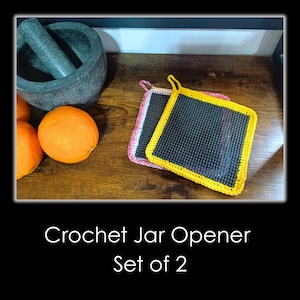 May include: Two crochet jar openers, one with a yellow border and the other with a pink border, are displayed on a wooden surface. The openers are square-shaped with a mesh centre. The text "Crochet Jar Opener Set of 2" is at the bottom.