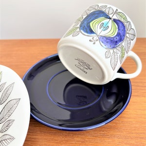 以下が含まれることがあります： 青と緑の花柄が描かれた白いセラミック製のコーヒーカップ。カップは逆さまになっており、ソーサーは濃い青色です。カップの底には「R&ouml;rstrand Sweden」の文字が見えます。