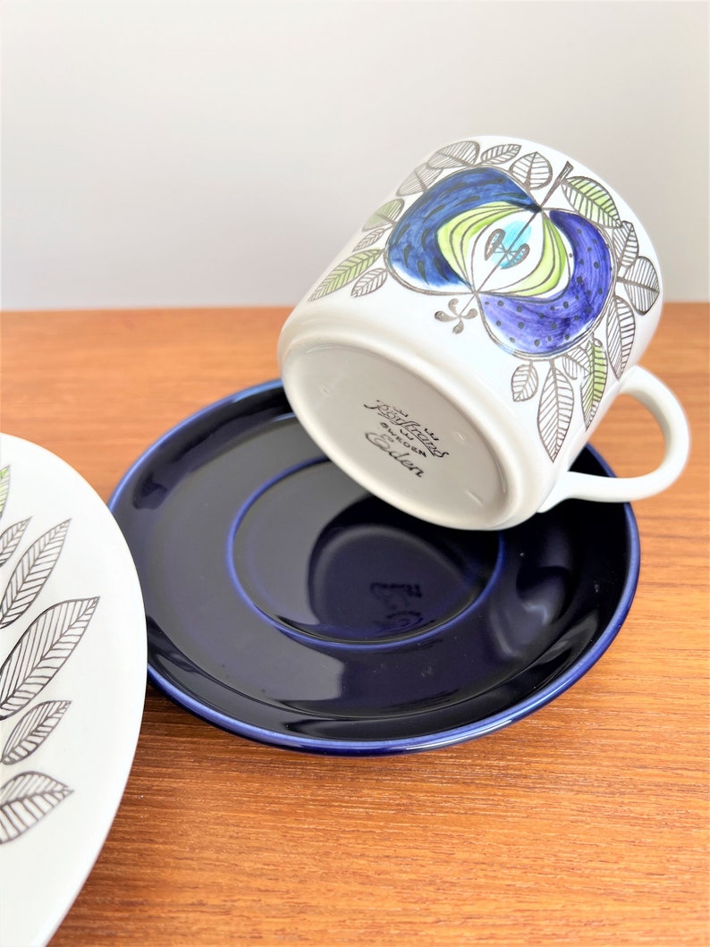 以下が含まれることがあります： 青と緑の花柄が描かれた白いセラミック製のコーヒーカップ。カップは逆さまになっており、ソーサーは濃い青色です。カップの底には「R&ouml;rstrand Sweden」の文字が見えます。