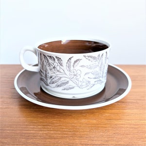 May include: A white teacup and saucer with a brown floral pattern. The cup has a brown rim and the saucer has a brown rim and a brown centre.