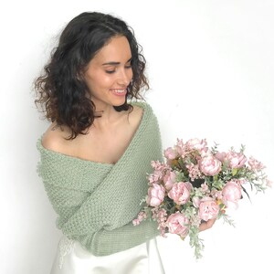 May include: A woman wearing a sage green knitted shrug with a white dress and holding a bouquet of pink peonies.