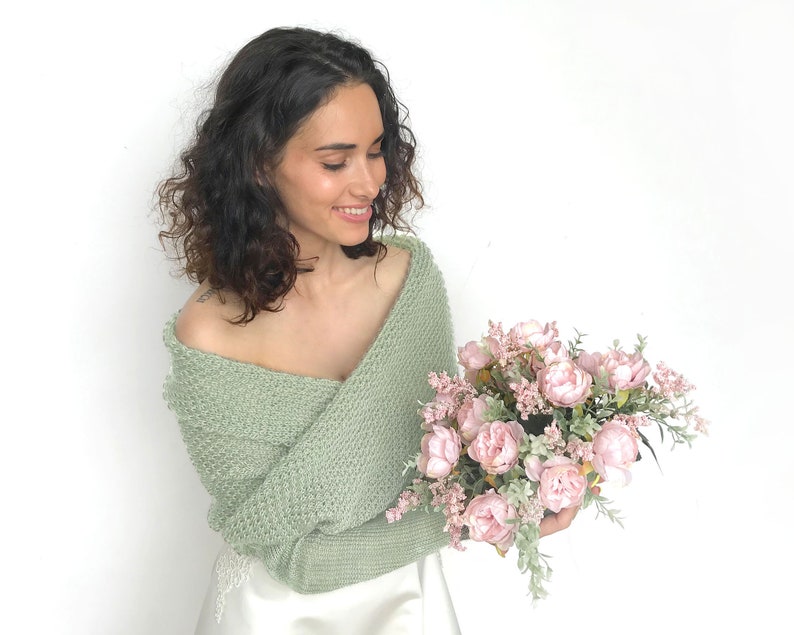 May include: A woman wearing a sage green knitted shrug with a white dress and holding a bouquet of pink peonies.