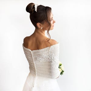 Peut inclure: Un boléro blanc en tricot à épaules dénudées, doté d'une rangée verticale de petits boutons ronds au milieu du dos et d'une broderie florale blanche complexe sur le côté droit, porté sur un vêtement blanc uni avec un accessoire de cheveux blanc.