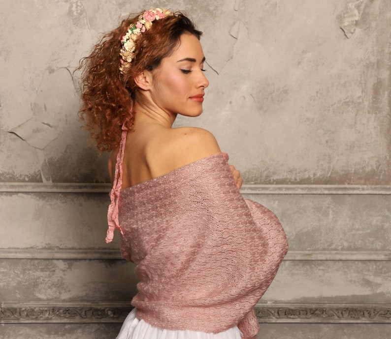 May include: A dusty rose knitted shawl with a textured, open-weave pattern draped over the shoulders. The garment features a soft, lightweight appearance and is paired with a floral headband adorned with small pink, cream, and green fabric flowers.