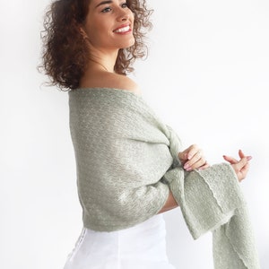 May include: A woman wearing a white dress with a sage green knitted shawl draped over her shoulders. She has curly brown hair with white flower hair clips.