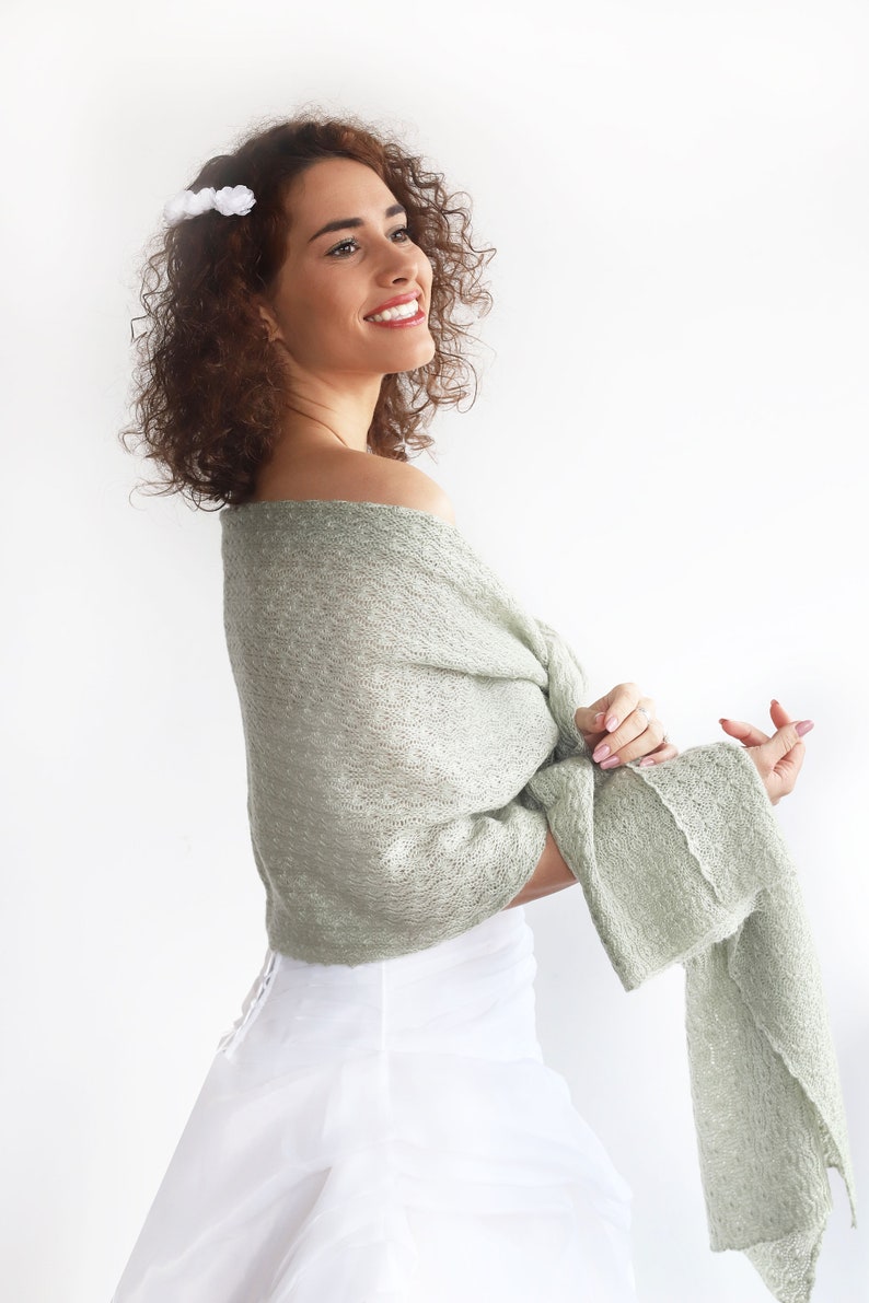 May include: A woman wearing a white dress with a sage green knitted shawl draped over her shoulders. She has curly brown hair with white flower hair clips.
