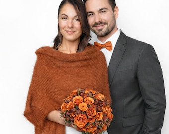 May include: A fuzzy, rust-colored knitted shawl draped over a white garment, paired with a bouquet of orange roses and dark berries. Beside it, a charcoal gray suit jacket is worn with a crisp white collared shirt and a matching rust-colored bow tie.