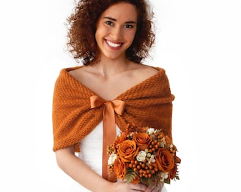 May include: A burnt orange knitted shawl featuring a textured stitch pattern and a matching satin ribbon tie closure at the center. The garment is draped over the shoulders and is paired with a bouquet of orange roses and small berries.