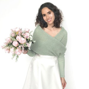 May include: A woman wearing a sage green knitted sweater with a white skirt and holding a bouquet of pink roses.