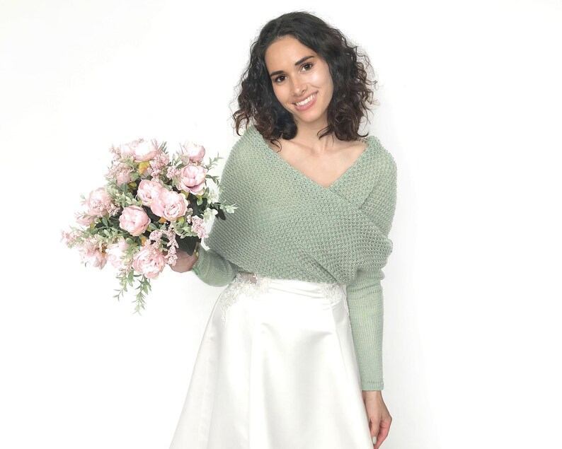 May include: A woman wearing a sage green knitted sweater with a white skirt and holding a bouquet of pink roses.
