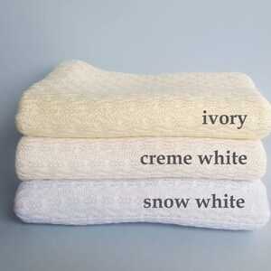 May include: A stack of three folded knitted blankets featuring a textured cable pattern. The top blanket is labeled ivory, the middle blanket is labeled creme white, and the bottom blanket is labeled snow white, with each color name printed next to it.
