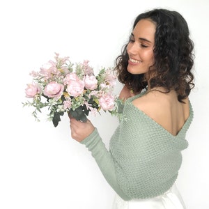 May include: A woman wearing a sage green knitted shrug with a low back, holding a bouquet of pink and white flowers.