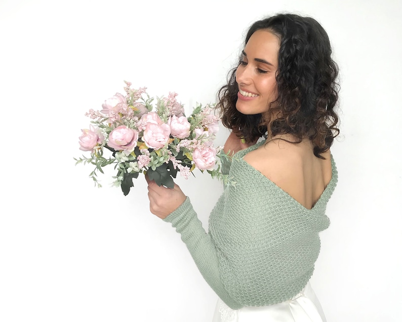 May include: A woman wearing a sage green knitted shrug with a low back, holding a bouquet of pink and white flowers.