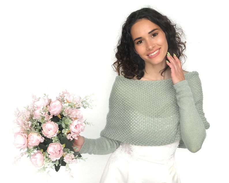 May include: A woman wearing a sage green knitted sweater with a bouquet of pink peonies and greenery.