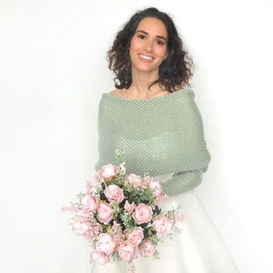 May include: A woman wearing a sage green knitted shawl over a white dress, holding a bouquet of pink roses.
