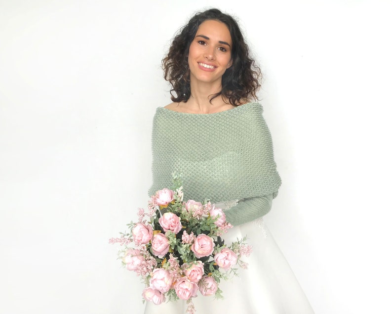 May include: A woman wearing a sage green knitted shawl over a white dress, holding a bouquet of pink roses.