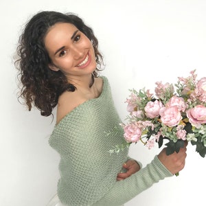 May include: A woman wearing a light green off-the-shoulder sweater holds a bouquet of pink peonies and greenery.
