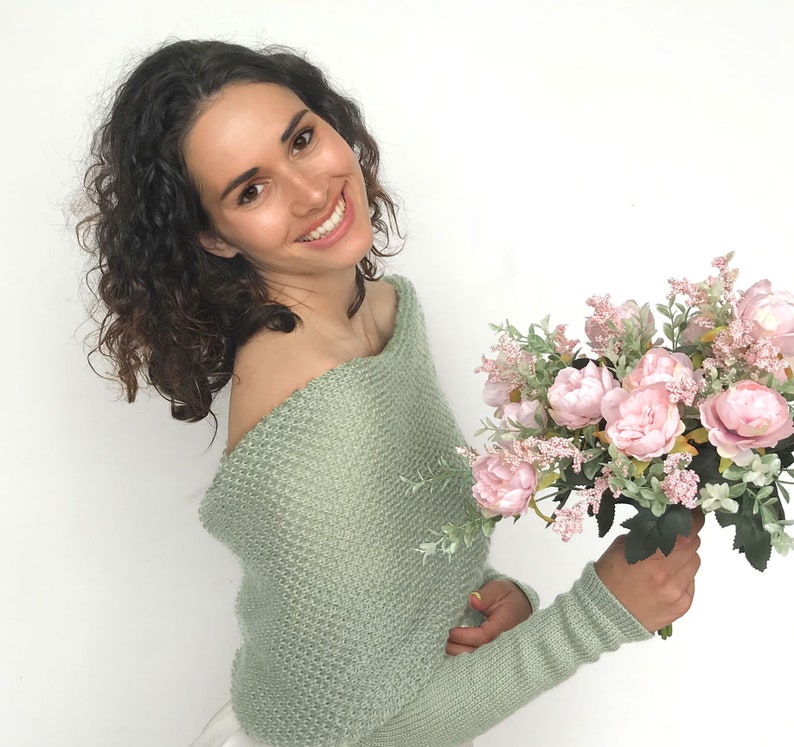 May include: A woman wearing a light green off-the-shoulder sweater holds a bouquet of pink peonies and greenery.