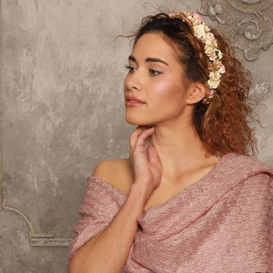 May include: A floral headband featuring small cream, white, and pink fabric flowers arranged in a dense cluster. The accessory is paired with a textured, dusty rose-colored knitted shawl that drapes over the shoulders with a visible open-weave pattern.