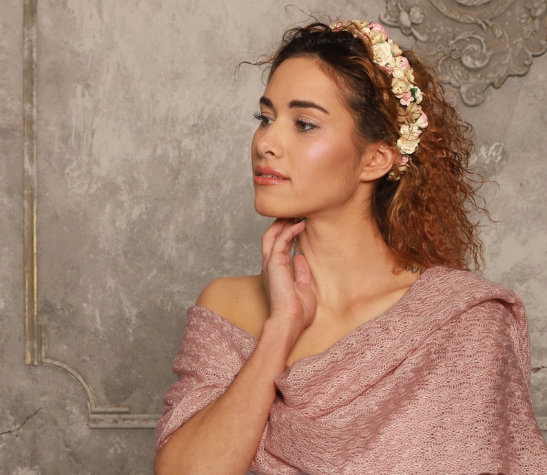 May include: A floral headband featuring small cream, white, and pink fabric flowers arranged in a dense cluster. The accessory is paired with a textured, dusty rose-colored knitted shawl that drapes over the shoulders with a visible open-weave pattern.