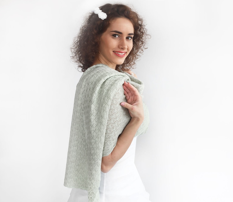 May include: A woman wearing a light green lace shawl with a white top. The shawl is draped over her shoulders and tied at the neck.