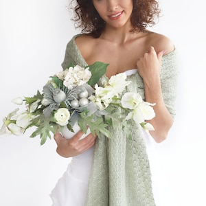May include: A woman in a white dress holds a bouquet of white flowers with silver accents. She is wearing a sage green shawl draped over her shoulders.