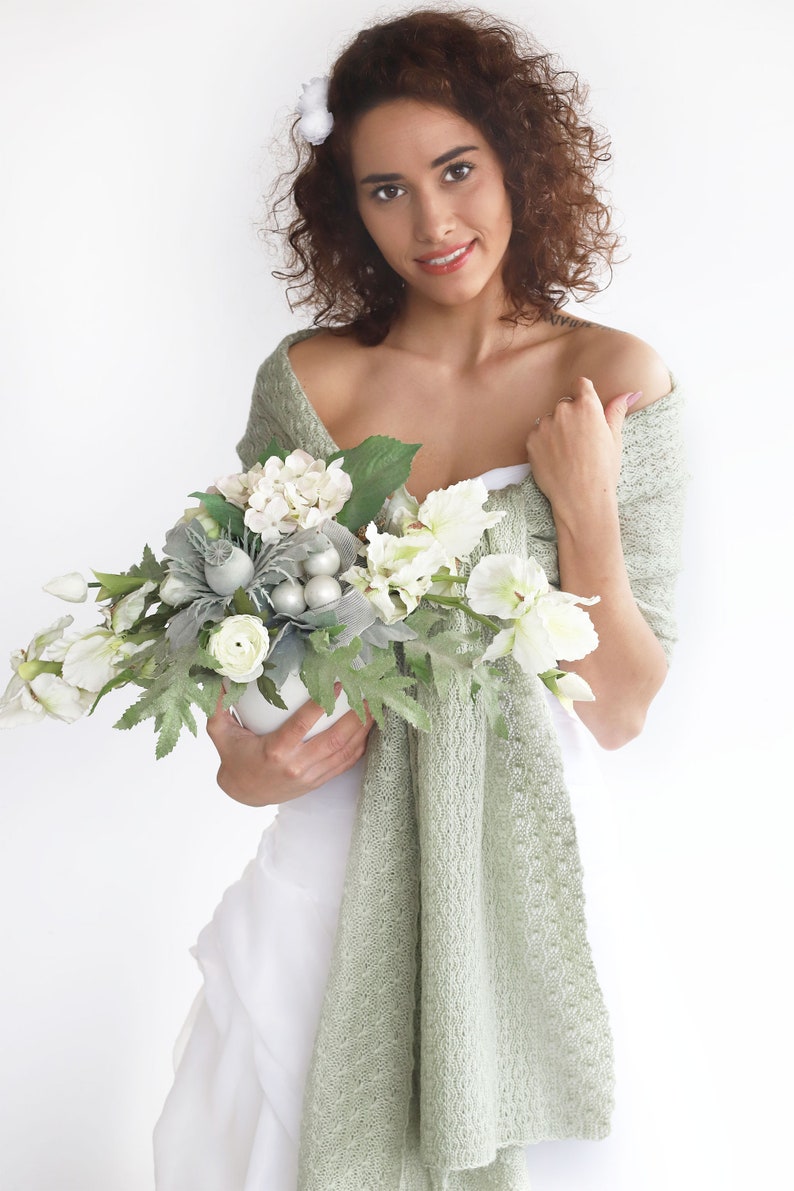 May include: A woman in a white dress holds a bouquet of white flowers with silver accents. She is wearing a sage green shawl draped over her shoulders.