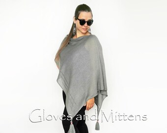 May include: Grey knitted poncho with a fringe detail. The poncho is being worn by a person with dark hair and sunglasses.