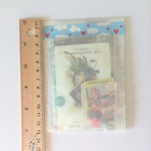 May include: A clear plastic bag containing vintage ephemera, including stamps, a postcard with floral imagery, and a small coin. The bag is sealed with a blue and white cloud-patterned tape with red hearts. A ruler is placed next to the bag.