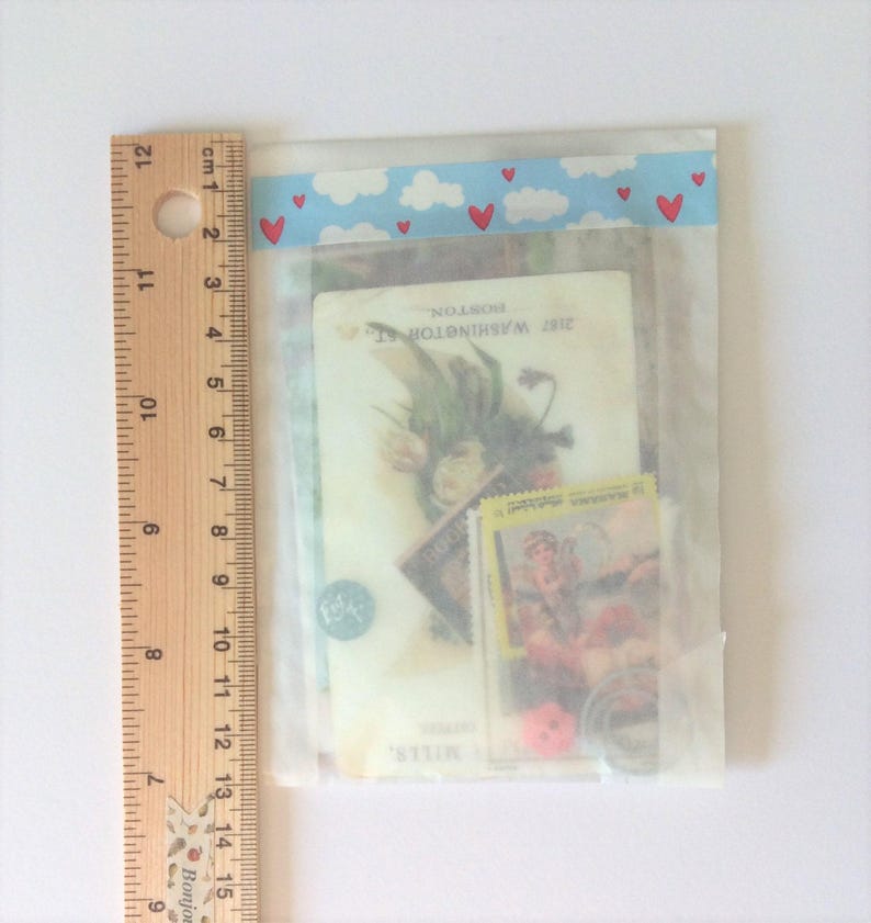May include: A clear plastic bag containing vintage ephemera, including stamps, a postcard with floral imagery, and a small coin. The bag is sealed with a blue and white cloud-patterned tape with red hearts. A ruler is placed next to the bag.