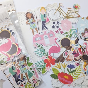 May include: A collection of paper stickers and die cuts featuring travel and vacation themes. The stickers include images of a pink flamingo, bicycles, a camper, and people. The color palette includes pink, green, gold, and white.