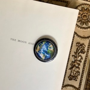 May include: A round button with a black border and a colourful image of the Earth against a dark blue background with white stars. The text "THE MOON AND" is visible in the background.