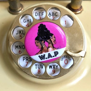 May include: A pink button with a graphic of two women with black hair and pink lips. The button has the text "W.A.P." in black letters on a white background. The button is on a rotary phone with gold colored buttons.
