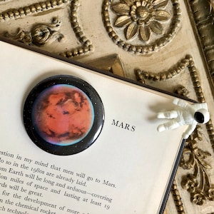 May include: A close-up of a round button featuring a colorful image of Mars, the red planet. The button is on a book with the word "MARS" printed on the cover. A small white astronaut figurine is on the book.