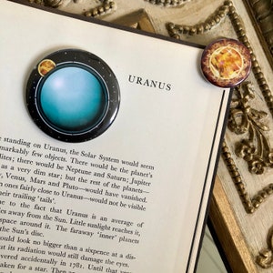 May include: A blue and black button with a graphic of the planet Uranus and its rings. The text "URANUS" is printed in black on a white background.